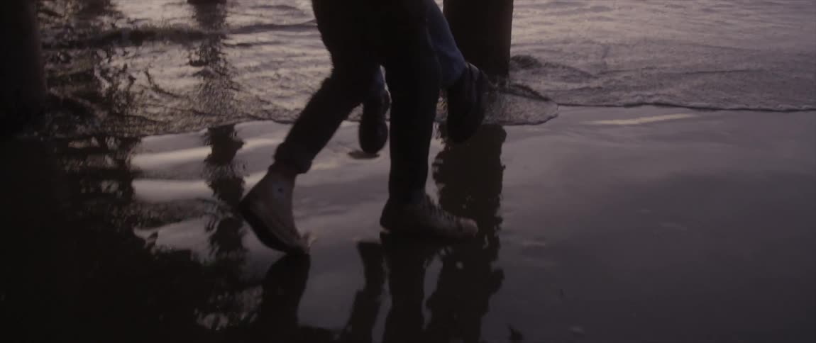 A man carries a woman through the water under a pier and puts her down on the beach sand