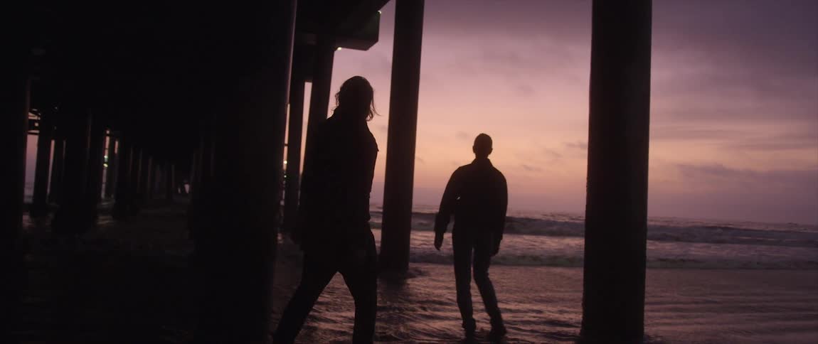 Silhouette of woman at the shoreline under pier at sunset as a man aggressively approaches
