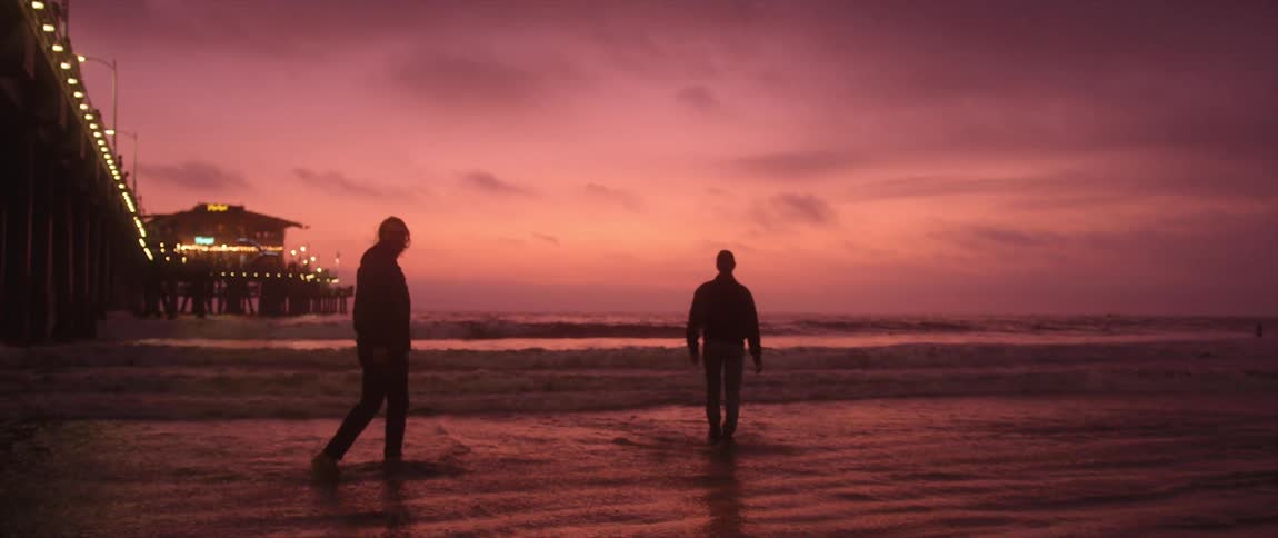 Two people walk in the water at the edge of the ocean near an illuminated pier at night