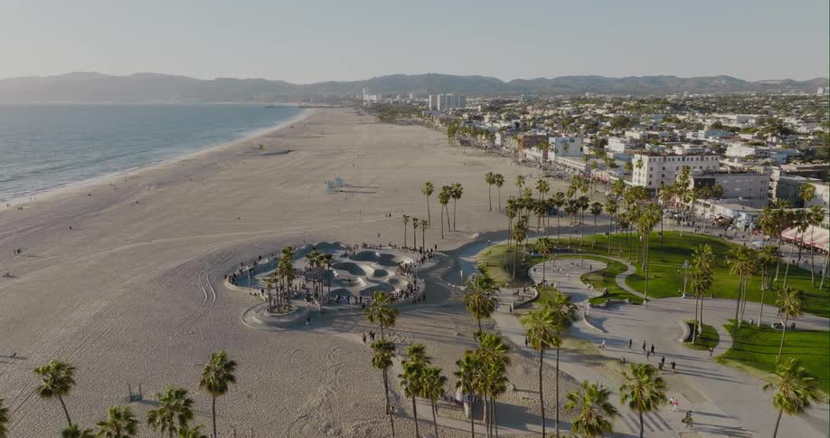 Pedestrians walking and people at a skateboard park on the beach next to the ocean as mountains sit in the distance