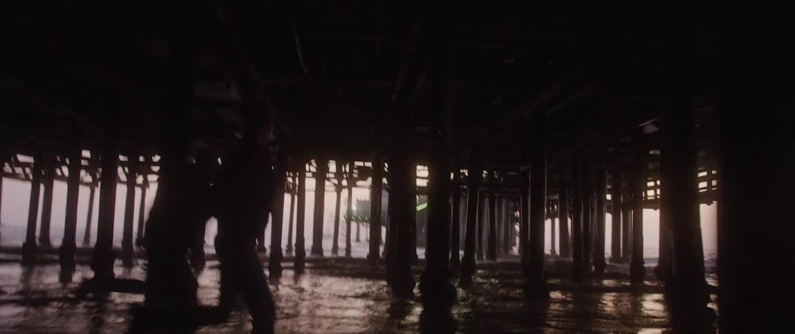 Two people run on the beach under a boardwalk