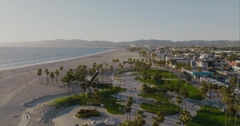 Ocean waves roll into shore as people skateboard at a skateboard park near the palm trees on the beach