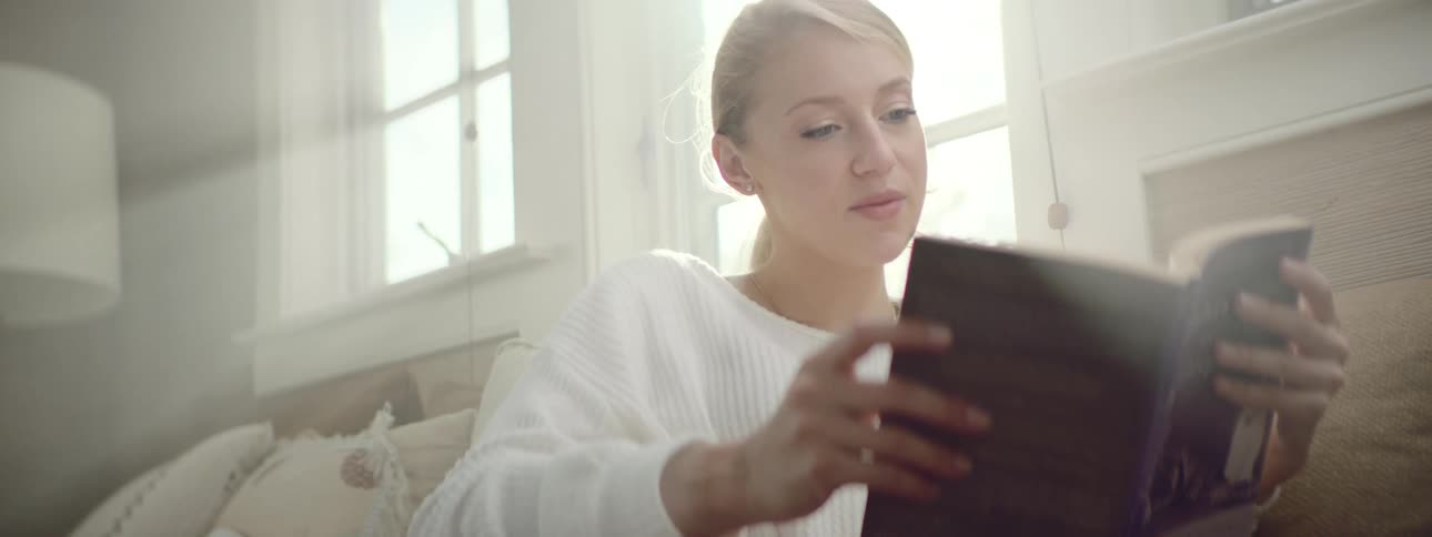 Young woman enjoys reading a book on the couch in a cozy living room with light streaming in