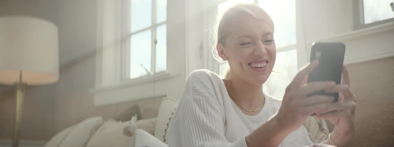 Woman sitting on the couch next to the window using her phone and laughing