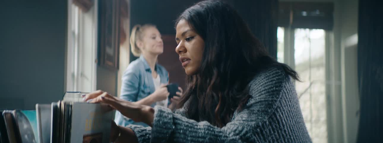 A woman sorts through records while her friend drinks coffee in a moody room