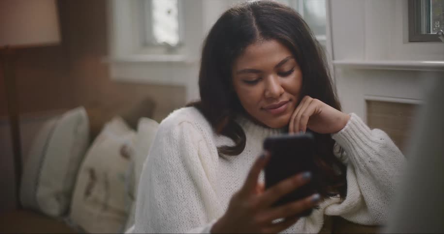 Woman with dark wavy hair and white sweater sits on couch and uses smartphone in living room at home