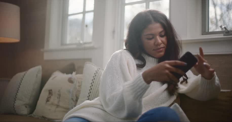 Young woman wearing a white sweater sits on a couch in the living room looking at her phone