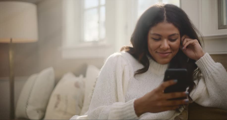 Woman with dark wavy hair and white sweater uses smartphone in living room at home