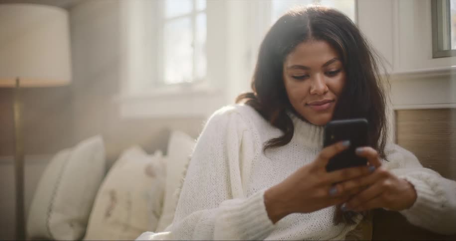 Woman with dark wavy hair and white sweater sits on couch using smartphone and smiling in living room at home