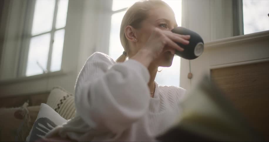 Blonde woman with ponytail and white sweater sits on couch and drinks from black coffee cup in living room at home