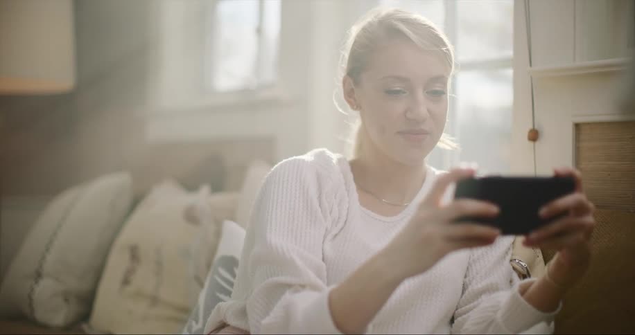A young woman sits at home and laughs as she looks at her cell phone