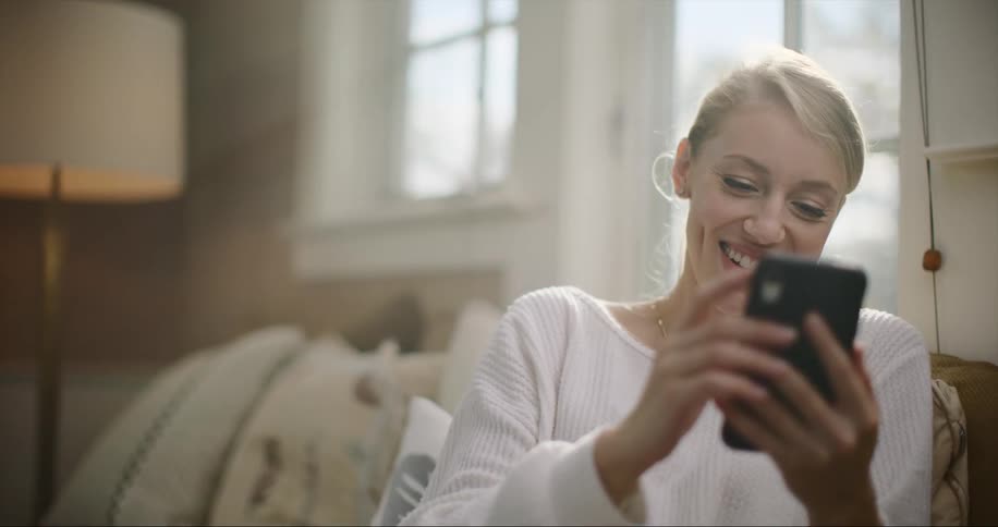 Blonde woman with ponytail and white sweater sits on couch using smartphone and smiling in living room at home