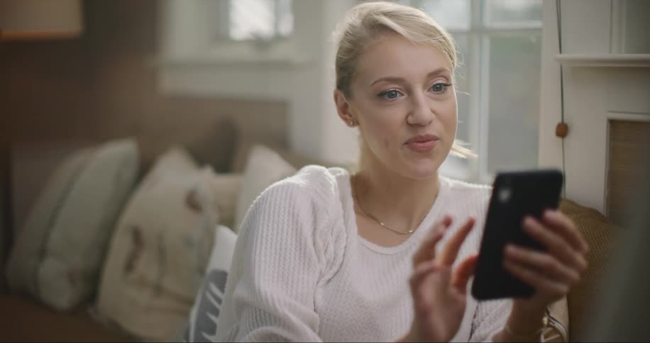 Blonde woman with ponytail sits on couch and uses smartphone in living room at home