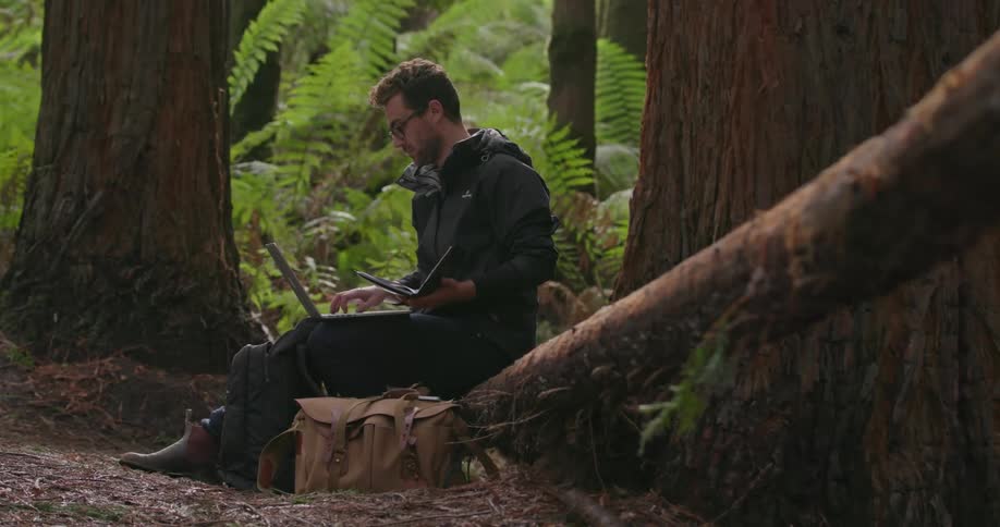 A man is sitting on a log with a laptop and a book