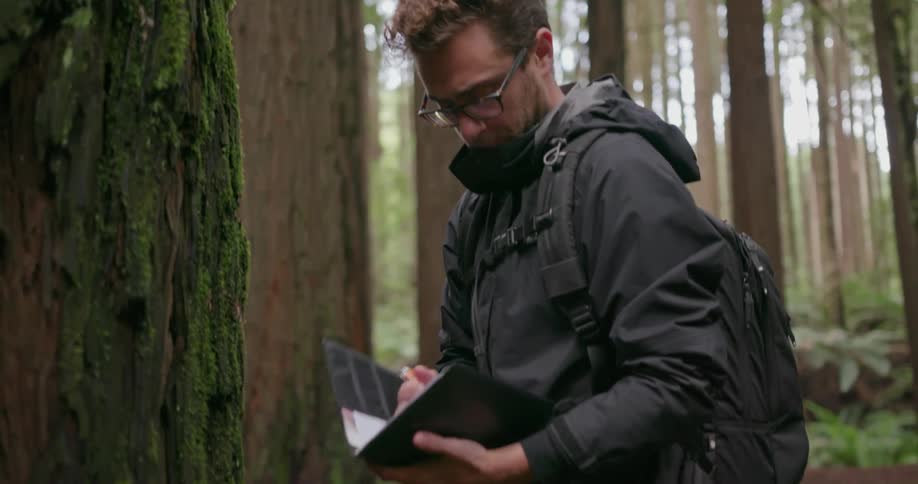 Man marking a tree with a pen and then writing in his book before moving on in a forest