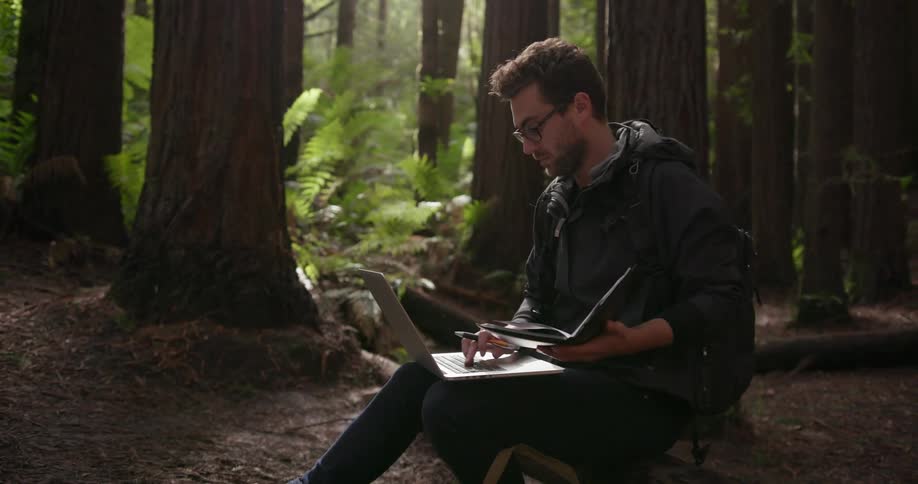 A man is using his laptop and writing in a notebook in the wilderness