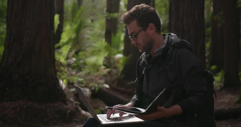 A man with a laptop and a book is sitting in the woods