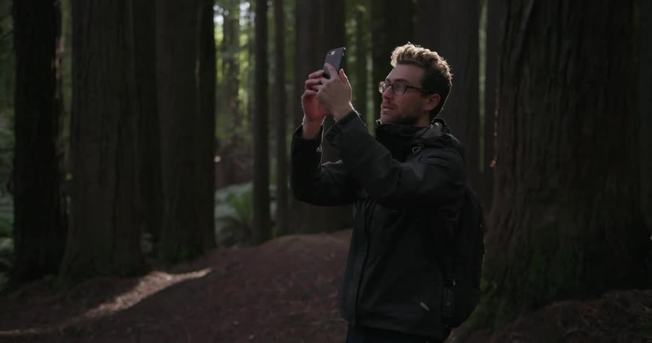 A man is taking pictures with his phone as he is hiking through the woods