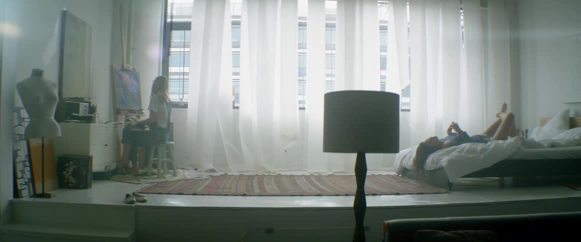 A woman is painting in the corner by a window as another woman lays down in bed