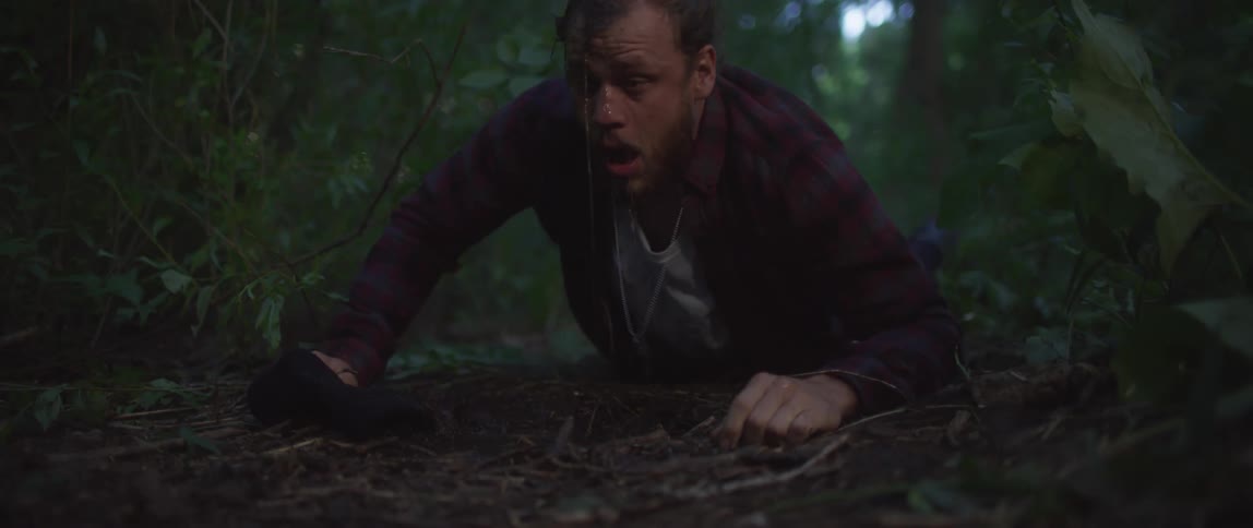 A man is lying on the ground on a trail in the woods as if he just woke up and he lifts his head and looks around