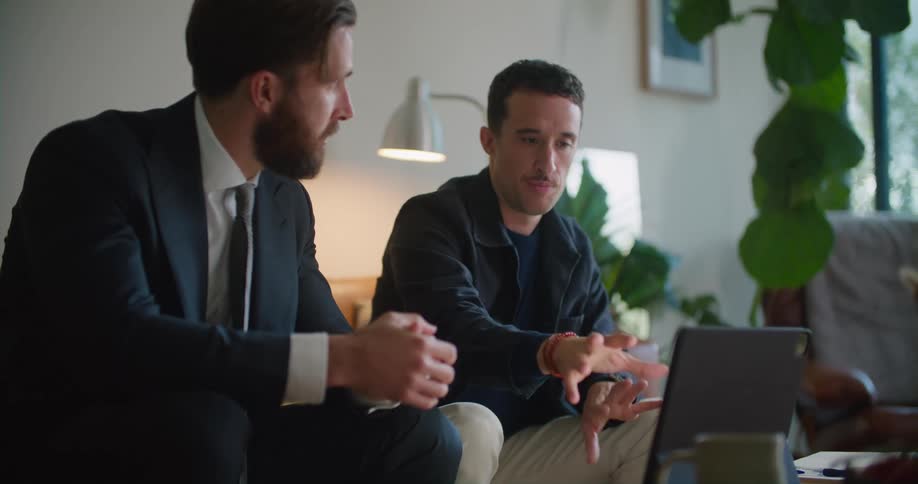 Two men sitting on the couch in the living room having a conversation in front of an open laptop at the center table
