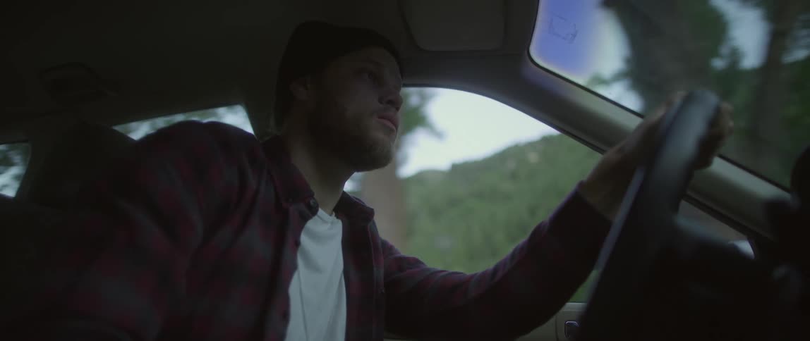 Young bearded man wearing a beanie drives a car near a mountain