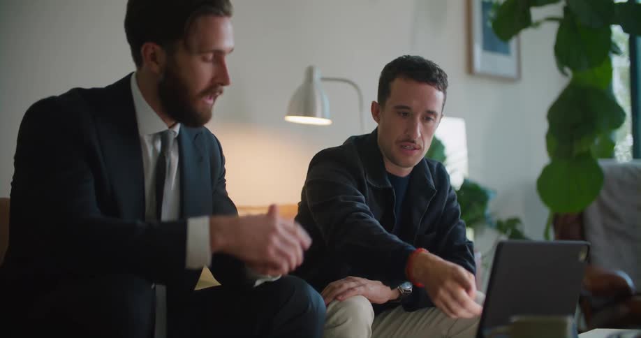 Business man wearing formal clothes sitting on the couch explaining something thats on the laptop screen to another man sitting next to him