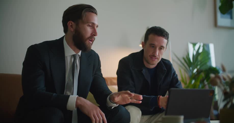 Man in business suit sits and talks to another man near laptop