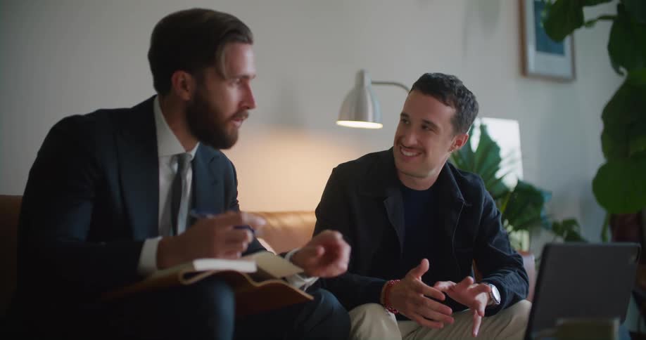 Man with mustache naval blue jacket shirt and beige pants sits on couch with man with suit and beard coming to an agreement in living room at home