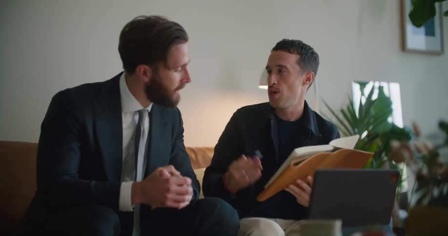 Man holds a pen and a book as he talks to another man sitting next to him