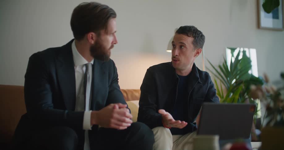 A pair of men talk with one another while sitting together on a sofa indoors during the day