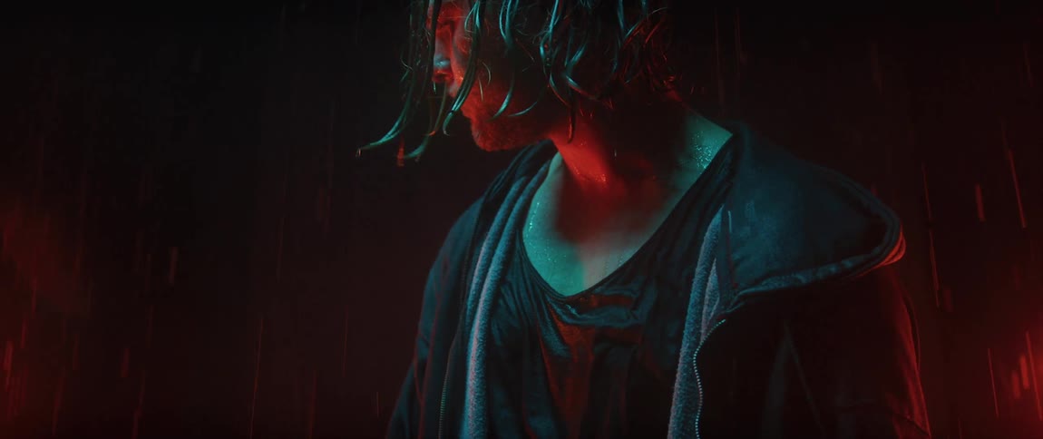 Man stands outdoors soaked in the rain under blue and red colored light