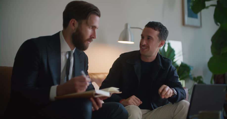 Business man sitting on the couch holding a notebook talking to a man next to him about something on the laptop screen