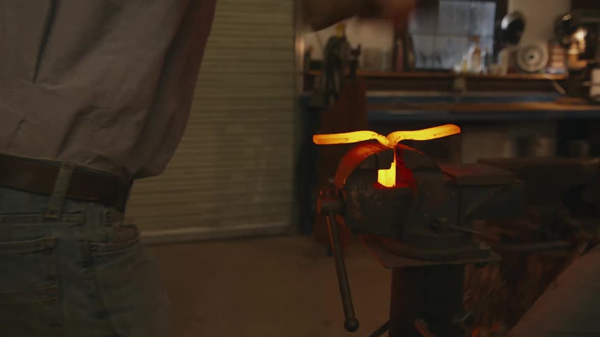 A man hammers pieces of metal glowing with heat in a vice with a mallet