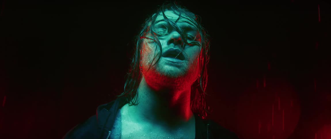 Man with long hair stands in red light while raining heavily in the street at night