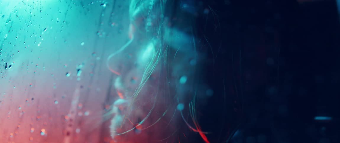 Young blonde man with long hair looking through a fogged window under neon lights at night