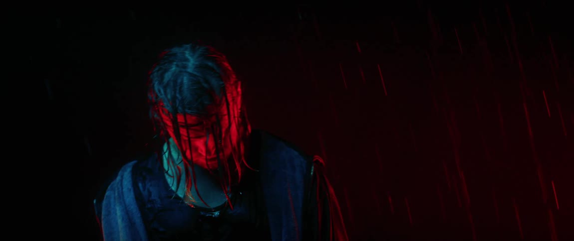 Man with long hair stands in red light while raining heavily in the street at night