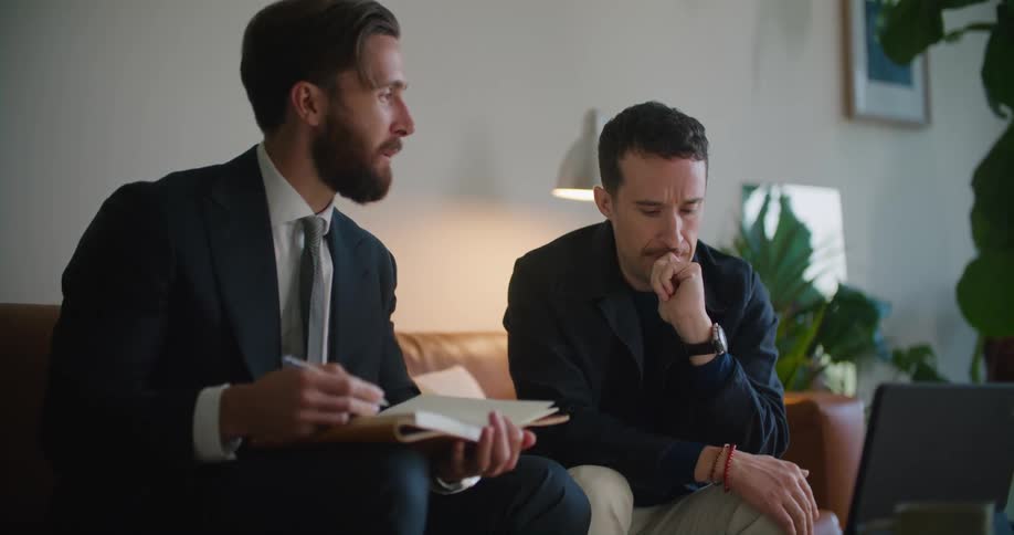 Business man wearing a suit sitting on the couch with a notebook on his lap talking to a man sitting next to him