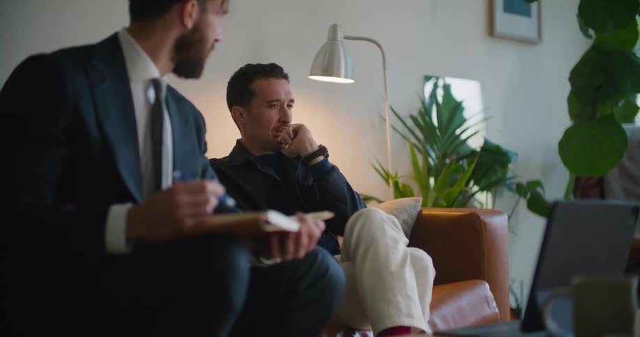 Two men sitting on the couch having a conversation and pointing at a laptop in front of them on the center table