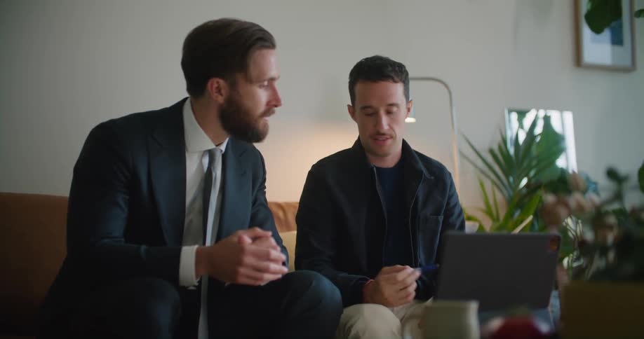 Two men sit on couch in office as one explains contents of book to the other