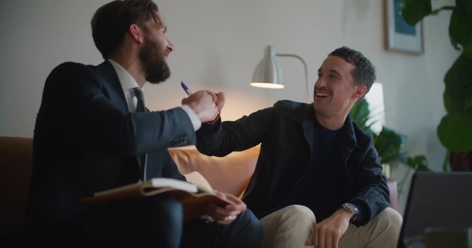 Two happy men sitting on the couch in a living room having a conversation while looking at a laptop screen at the center table