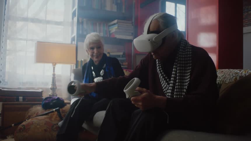 An elderly man puts on a virtual reality headset as a woman encourages him while he swings his arms but he takes it off and gives it to her to try