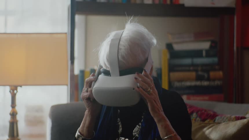 Elderly woman with gray hair navy blue shirt wears virtual reality headset sitting on couch in living room