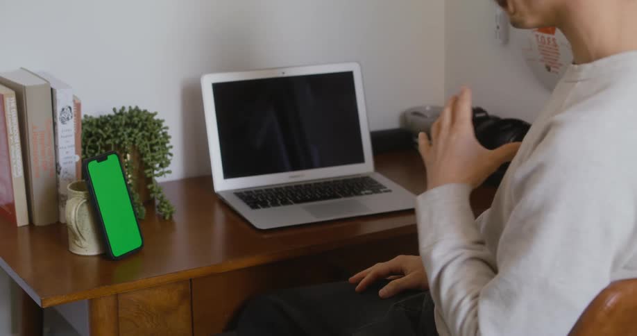 Man with white sweatshirt props smartphone against cup on table and talks to smartphone