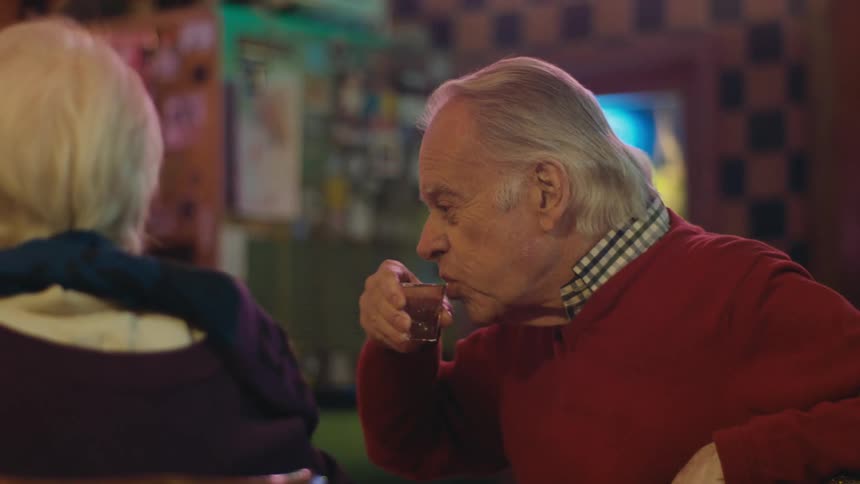 An elderly man talks to the woman sitting next to him in a diner as he drinks from a shot glass