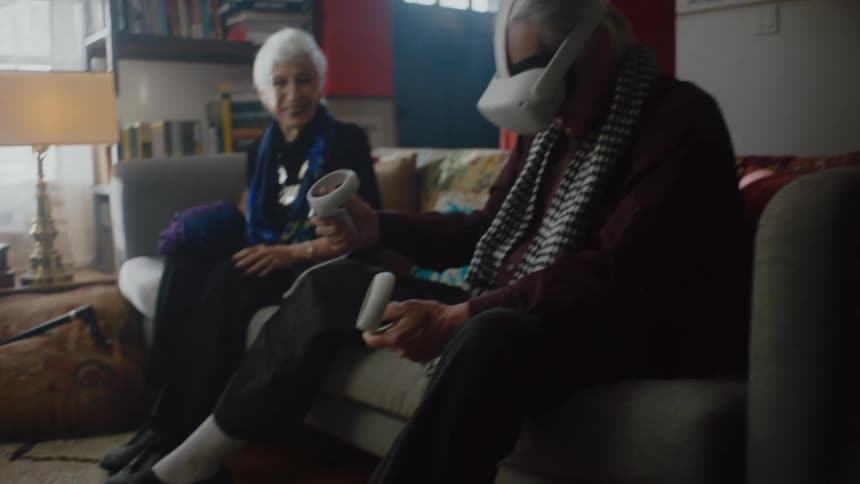 Elderly man sitting on couch uses virtual reality headset next to elderly woman in living room