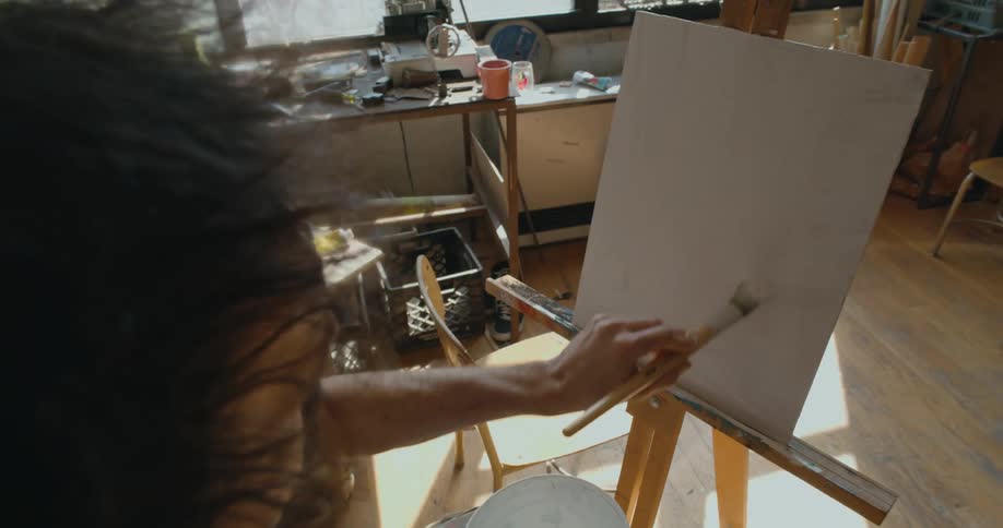 Painter sitting in front of a canvas panel on an easel uses a brush to spread white paint