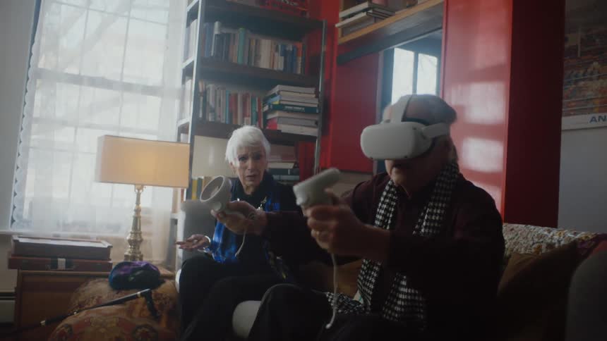 An elderly man plays a game using a virtual reality headset while an elderly woman sitting beside him on a sofa watches