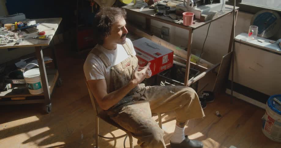 Man sitting in chair in paint splattered overalls uses sign language
