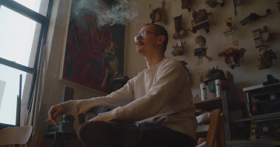 A man sits and smokes a cigarette next a window at home